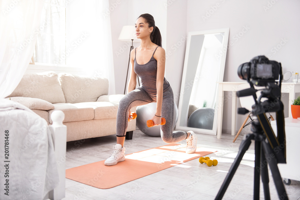 Fototapeta premium Exercising tutorial. Fit young woman doing lunges and holding dumbbells while recording herself on complex