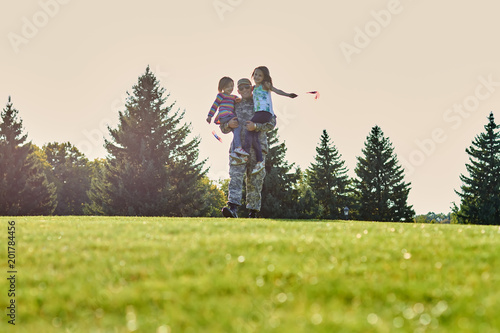 Portrait of soldier is holidng his daughters. Father im military uniform is walking with two little kids on the grass.