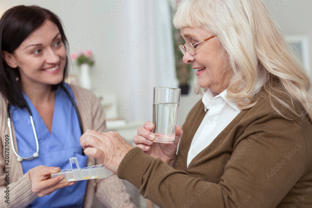 Managing medication. Positive mature woman taking pills from nurse while looking down Managing medication. Positive mature woman taking pills from nurse while looking down