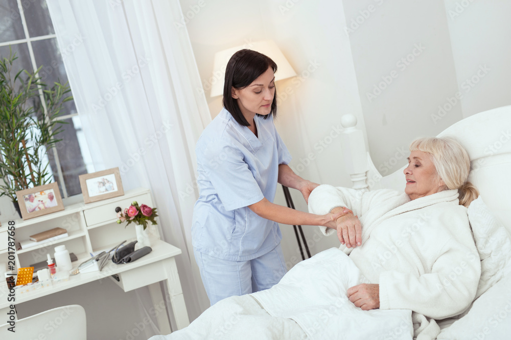 Fototapeta premium Little effort. Concentrated appealing nurse helping elder woman who lying in bed