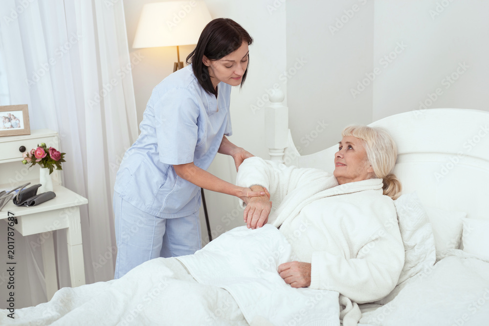 Stay calm. Pleased experienced nurse supporting elder woman who smiling in bed