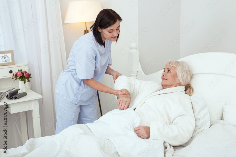 Fototapeta premium Stay calm. Pleased experienced nurse supporting elder woman who smiling in bed