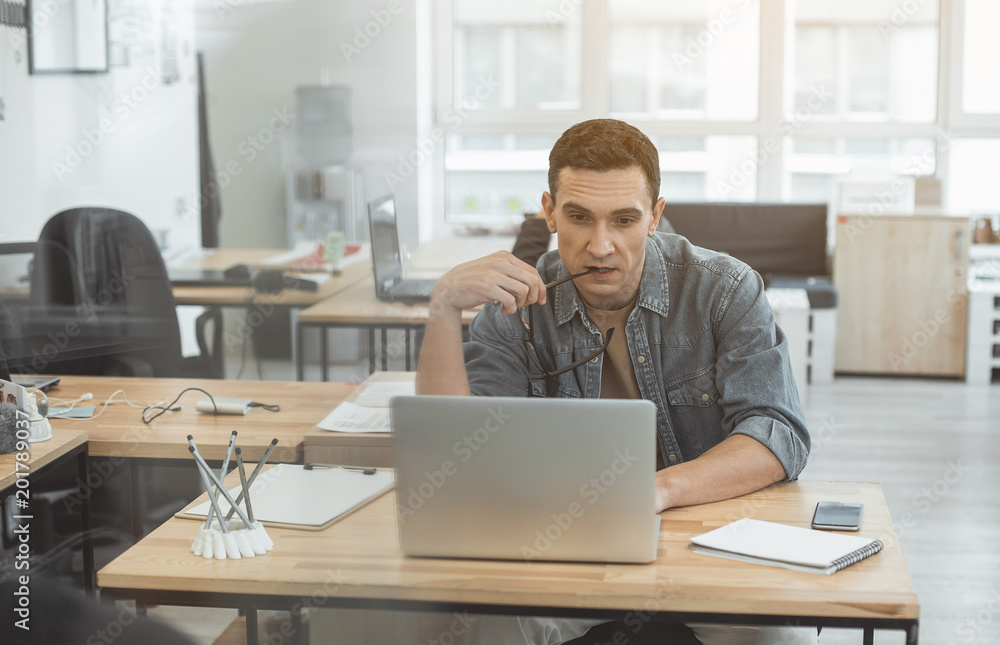 Portrait of thoughtful man looking at laptop while locating at desk in office. Wistful employee during job with gadget concept Portrait of thoughtful man looking at laptop while locating at desk in office. Wistful employee during job with gadget concept