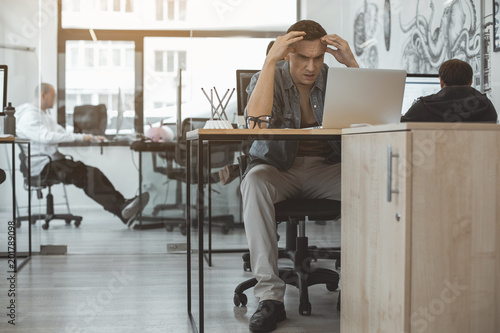 Full length portrait of undeceived man looking at notebook computer while sitting at table. Disillusion during job concept photo