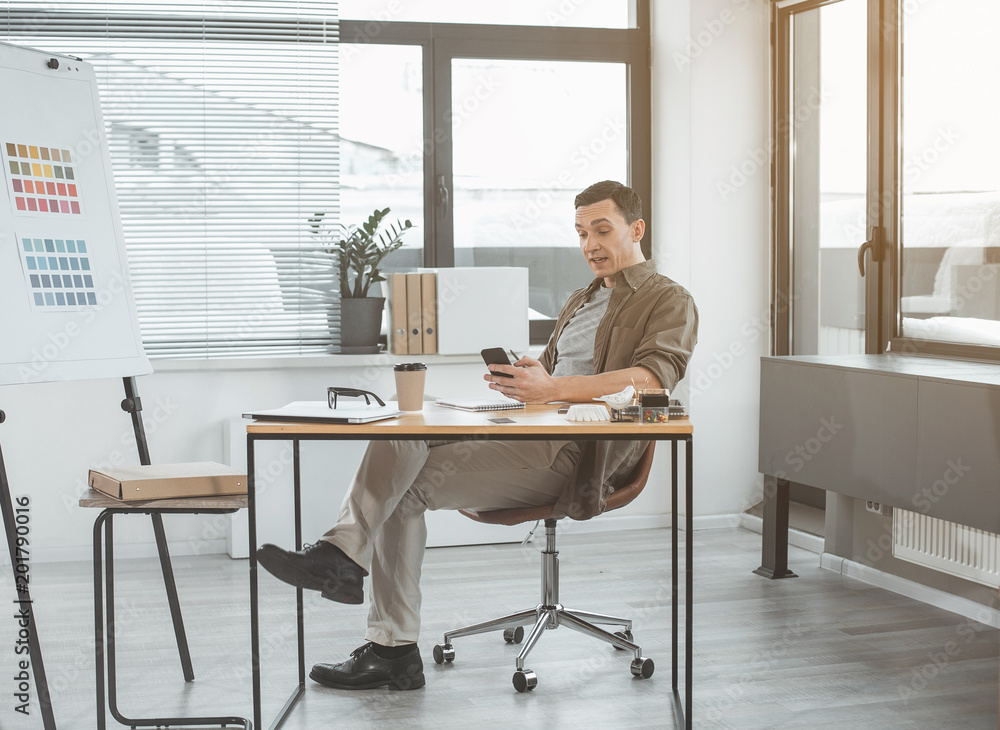 Full length portrait of surprised designer using contemporary mobile while locating at table during labor. Shocked employer having job with technology concept