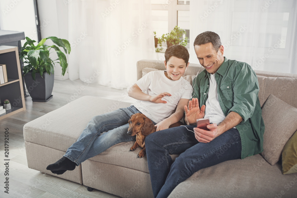 Joyful man and boy speaking online while waving hands to smartphone camera. They are relaxing on sofa with dachshund pet and laughing Joyful man and boy speaking online while waving hands to smartphone camera. They are relaxing on sofa with dachshund pet and laughing