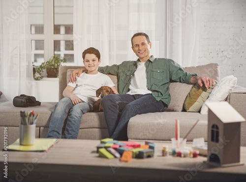 Portrait of happy father and son are resting on couch together with dog. They are looking at camera and smiling