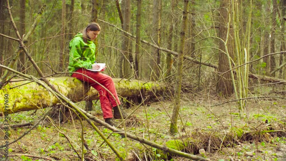 Woman sitting on the tree in a spring forest and writing something in notebook. The scientist ecologist during the research. Woman sitting on the tree in a spring forest and writing something in notebook. The scientist ecologist during the research.