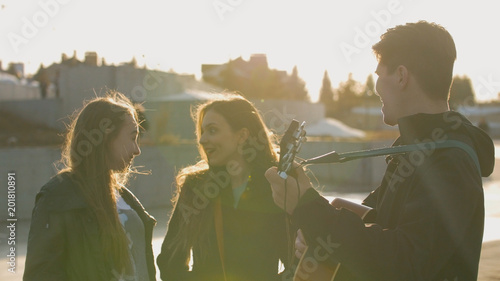 Two girls smiling while guy playing on the guitar at the sunset outdoors