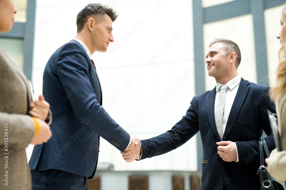 Content confident handsome young businessmen in formal suits making handshake after signing contract in office Content confident handsome young businessmen in formal suits making handshake after signing contract in office