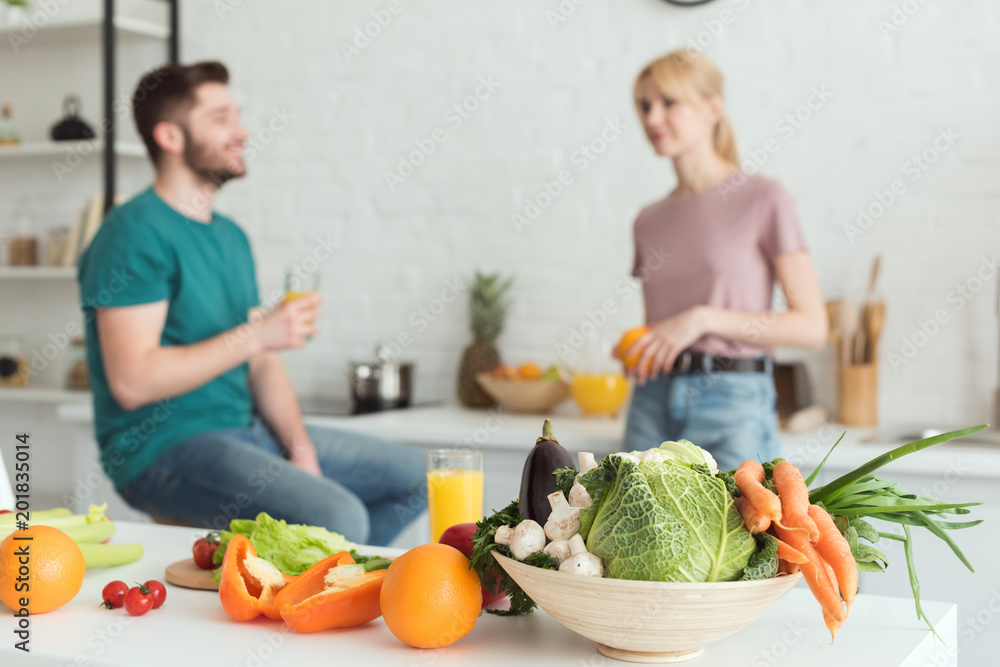 vegan couple talking in kitchen with fruits and vegetables on foreground vegan couple talking in kitchen with fruits and vegetables on foreground