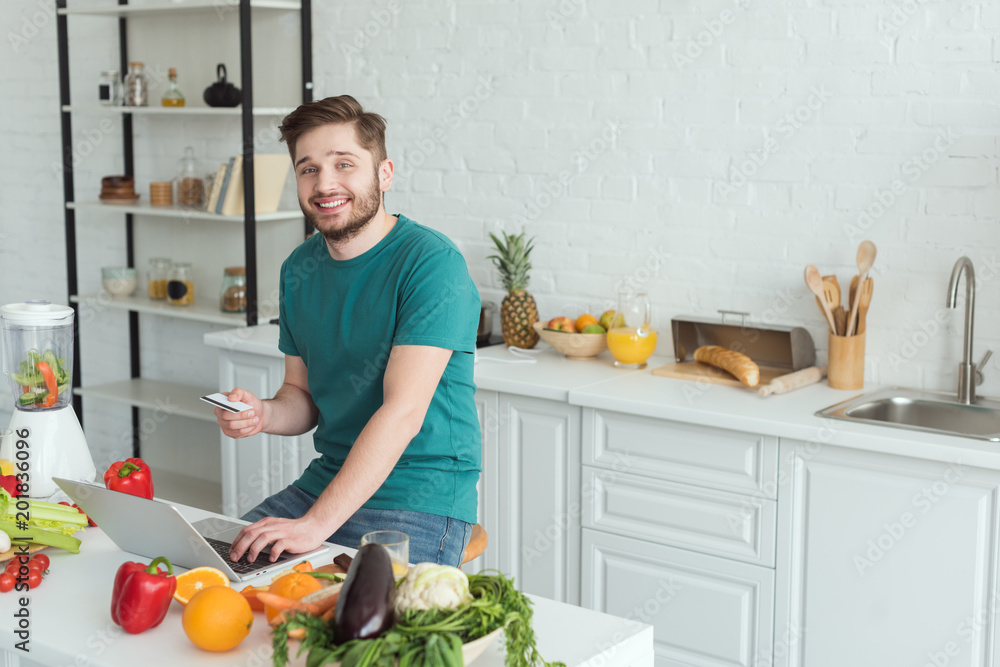 smiling man with credit card sitting at table with laptop in kitchen at home smiling man with credit card sitting at table with laptop in kitchen at home