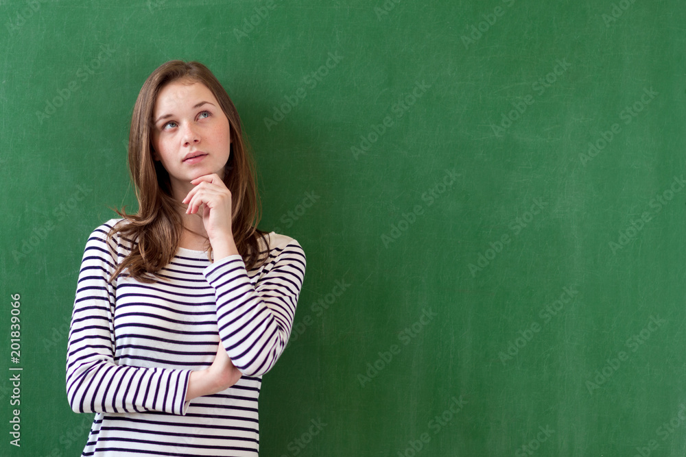 Student thinking and leaning against green chalkboard background. Pensive girl looking up. Caucasian female student portrait with copy space. Imagination, ideas, future, possibilities concept. Student thinking and leaning against green chalkboard background. Pensive girl looking up. Caucasian female student portrait with copy space. Imagination, ideas, future, possibilities concept.