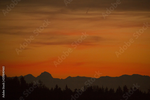 Orange silhoue tte of a dark horizon with a silhoutte of a mountain range of the alps on a sunny day evening after sunset with clouds