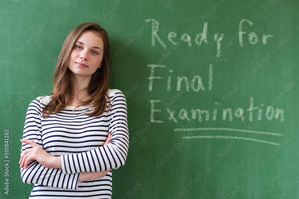 Young confident smiling female high school student standing in front of chalkboard in classroom, with her arms crossed, looking at camera. Ready for exams concept.
