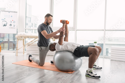 Fototapeta Naklejka Na Ścianę i Meble -  Rehabilitation training. Pleasant handsome man lying on the medball while lifting dumbbells