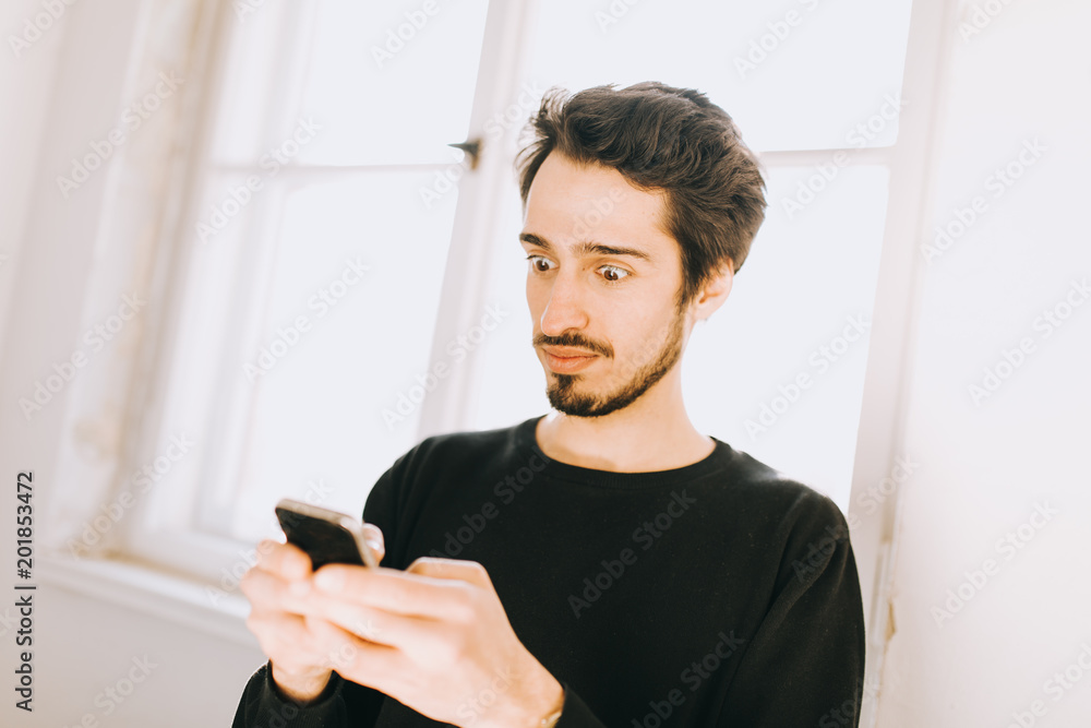Junger dunkelhaariger Mann schaut erstaunt auf sein Handy Junger dunkelhaariger Mann schaut erstaunt auf sein Handy