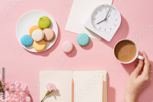 Female hand with coffee cup, macaron, office supply, gift and notebook on pastel desk top view. Fashion pink woman workplace background for blogging. Flat lay. Beautiful morning breakfast.