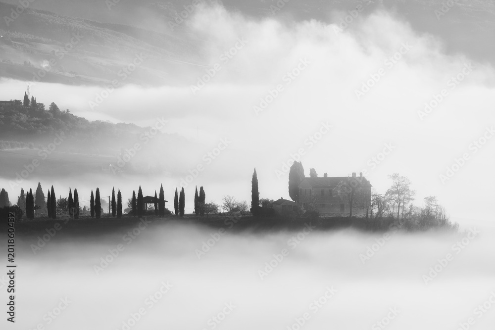 Beautiful foggy sunrise in Tuscany, Italy with cypresses and house. Natural misty background in black and white
