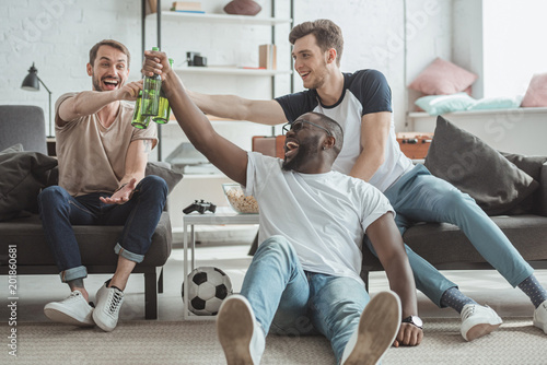 multiethnic young men watching football match and celebrating and clinking bottles of beer