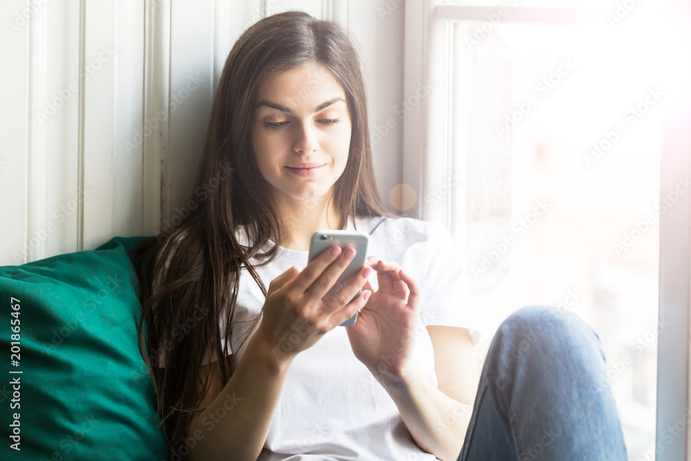 Beautiful smiling brunette girl wears white t-shirt using smartphone sitting on the windwsill Beautiful smiling brunette girl wears white t-shirt using smartphone sitting on the windwsill