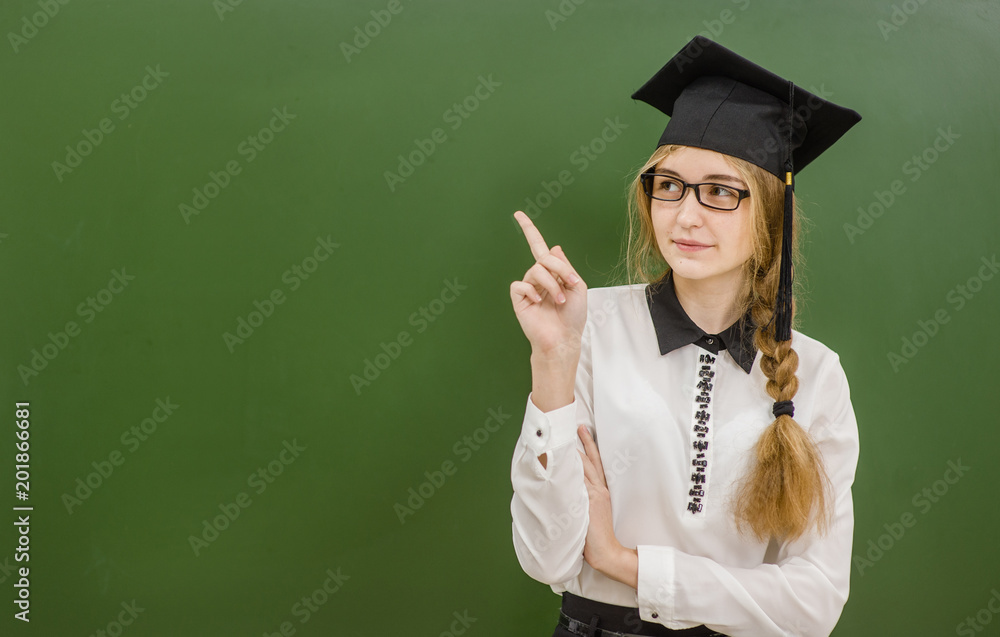 Teen girl in graduation cap standing near green chalkboard and pointing on empty space Teen girl in graduation cap standing near green chalkboard and pointing on empty space