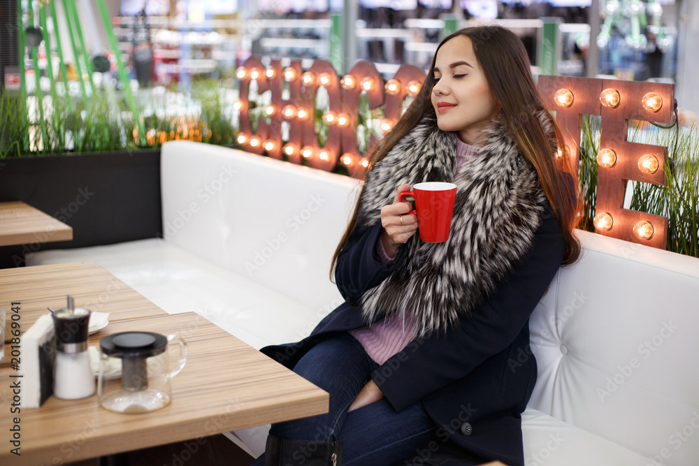 Portrait of a beautiful brunette girl in a winter coat with a cup in hands sitting in a cafe.