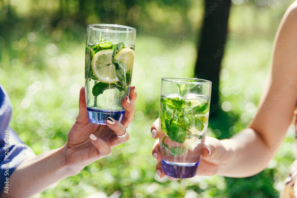 Two female hands holding glasses filled water with lemon and green mint leaves. Picnic in the garden. Detox, healthy eating, drinks, diet and people concept. Summer, outdoors.