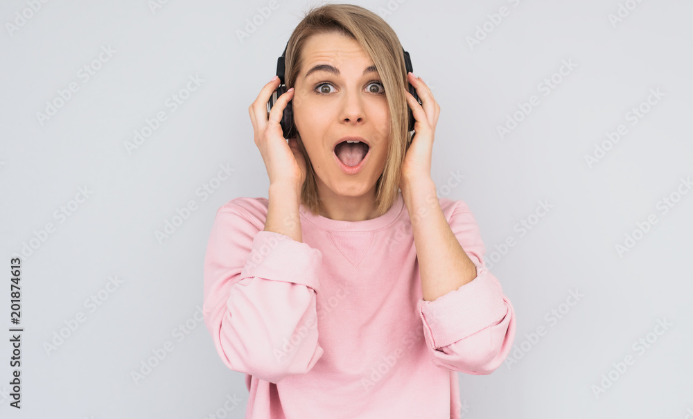 Studio portrait of amazed young female wears pink sweater looking at the camera, holding headphones with hands, listening favorite music, isolated on white background. Copy space for advertisement Studio portrait of amazed young female wears pink sweater looking at the camera, holding headphones with hands, listening favorite music, isolated on white background. Copy space for advertisement