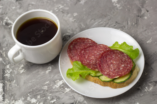 cup of black coffee and sandwich with sausage, cucumber and salad