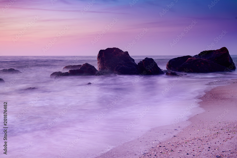 Summer seasonal natural vacation background. Romantic morning at sea. Big boulders sticking out from smooth wavy sea. Pink horizon with first hot sun rays. Long exposure.