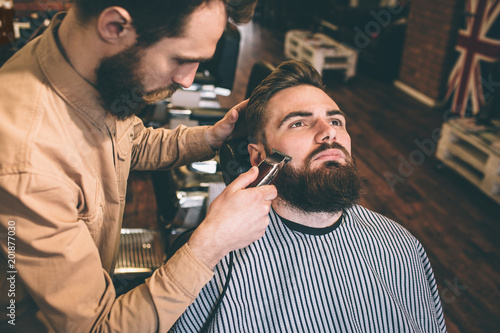 Nice and good hairstylist is cutting some of the customer's beard with an electrical razer. He is doing that very accurate.