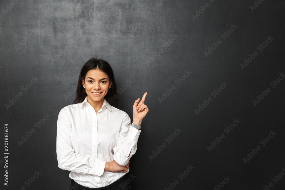 Portrait of secretary or business woman with long dark hair wearing white shirt pointing finger upward on copy space, isolated over dark gray background Portrait of secretary or business woman with long dark hair wearing white shirt pointing finger upward on copy space, isolated over dark gray background