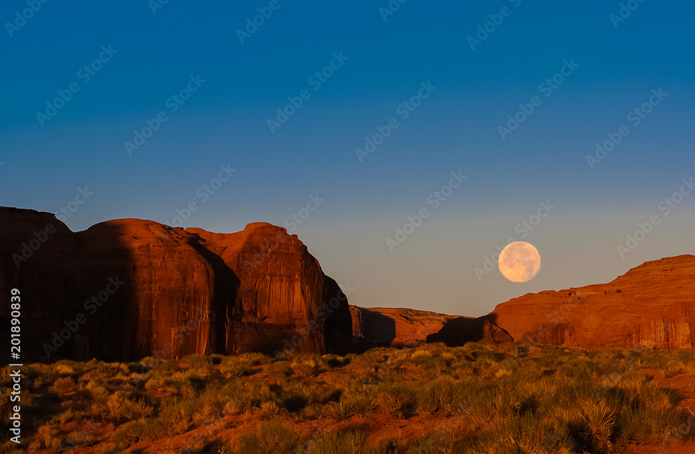 Morning landscape in Monument Valley Navajo Tribal Park with moon setting down while the rising sun is enlightening red sandstone cliffs.