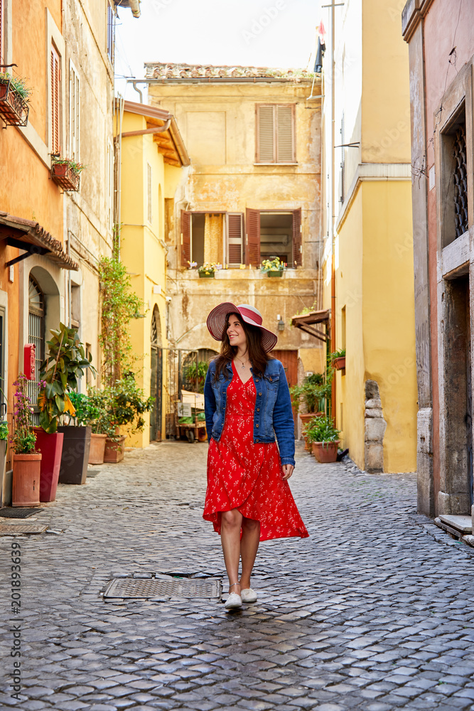 Happy woman in dress and hat walking on paved old street of Rome looking happily around.