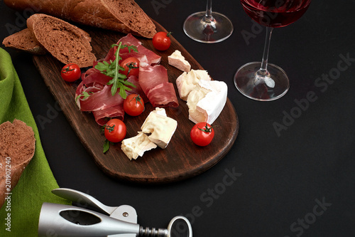 Cheese, prosciutto, bread, vegetables and spices on wooden board on black background with copy space