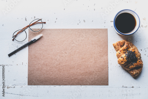 Mockup blank craft sheet of blank craft paper, pen, eye glasses and morning coffee cup with croissant on white wooden desk. Business clean mock-up background for message writing.Creative work concept photo