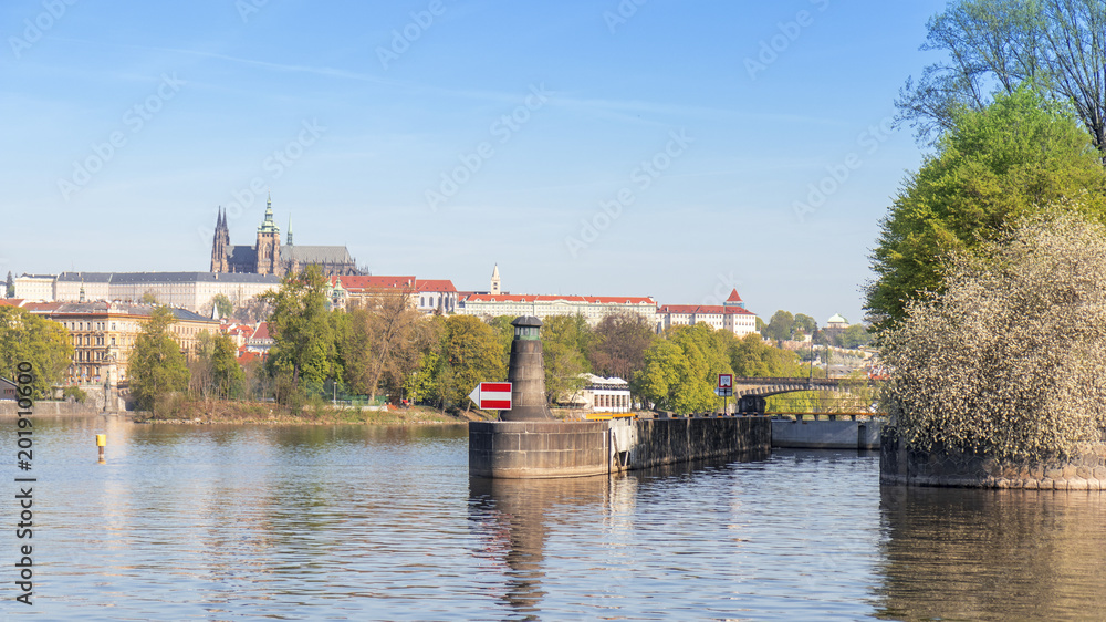 Obraz premium The lighthouse, navigation or orientation landmark and the lock, gate built on the riverbank of one of Prague island Zofin. The cityscape of Prague Castle in the background of image.