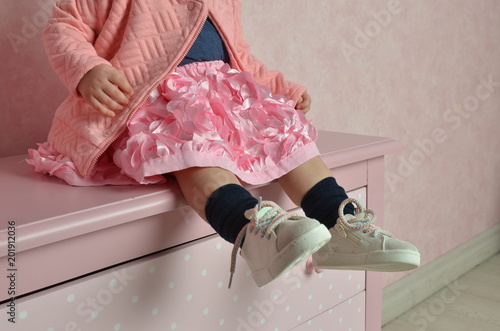 Legs of a little girl sitting on a dresser. close-up shot of the baby's legs in a pink skirt and white sneakers. Stylish kids. Pink photo