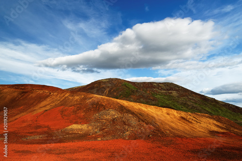 Colorful deposits of volcanic ash in reds and yellows against a green hill, above blue sky with a striking cloud formation - Location: Iceland, Golden Circle