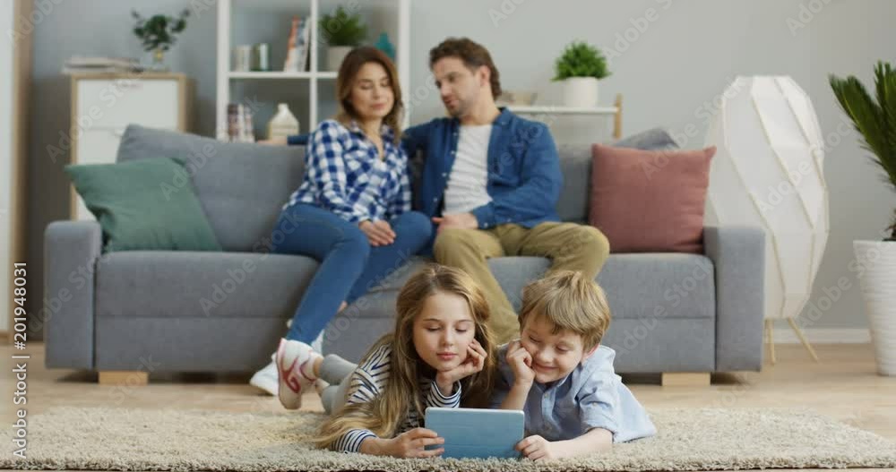 Two pretty cute kids, son and daughter, lying on the carpet in the living room and watching something funny on the tablet device. Their parents sitting on the couch behind them. Indoors Two pretty cute kids, son and daughter, lying on the carpet in the living room and watching something funny on the tablet device. Their parents sitting on the couch behind them. Indoors
