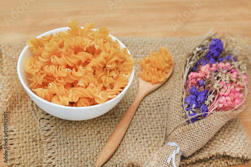 Spiral Macaroni in white bowl on sack decoration with dry flower bush against wood background.