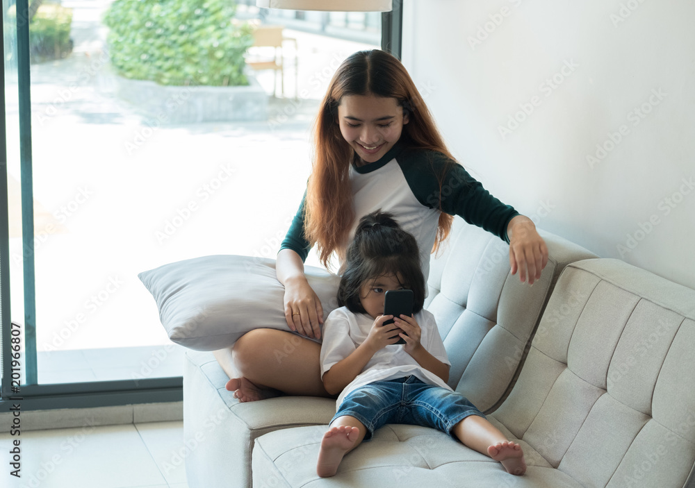 Two sisters use mobile phone and sitting at sofa in house with relax emotion,Family lifestyle concept. Two sisters use mobile phone and sitting at sofa in house with relax emotion,Family lifestyle concept.