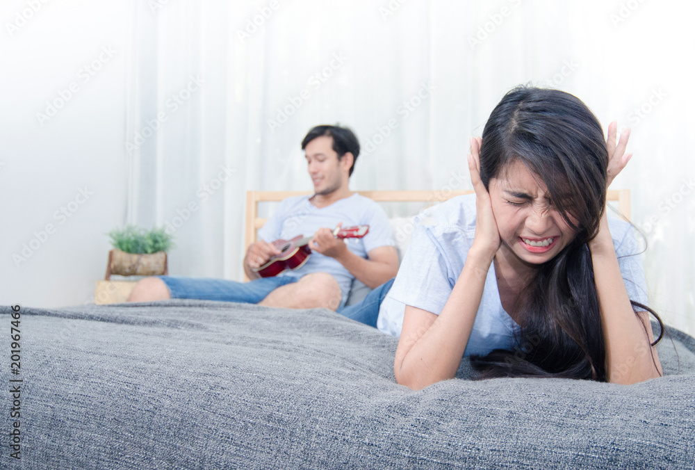 Woman feeling annoyed and stressed on the bed with man play ukulele in background Woman feeling annoyed and stressed on the bed with man play ukulele in background