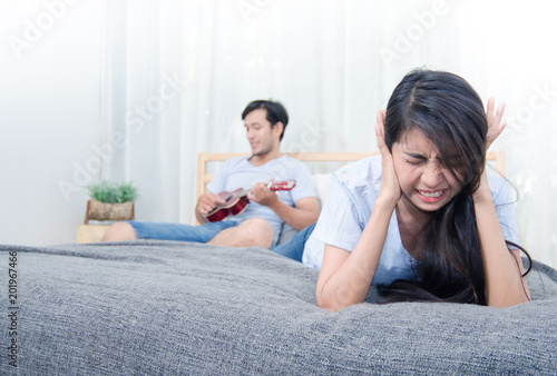 Woman feeling annoyed and stressed on the bed with man play ukulele in background