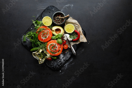 Delicious Salmon rolls, goats cheese in paper with knife, aromatic herbs, spices  on old black stone background, clean eating, healthy food, diet or cooking concept, top view, overhead, flat lay