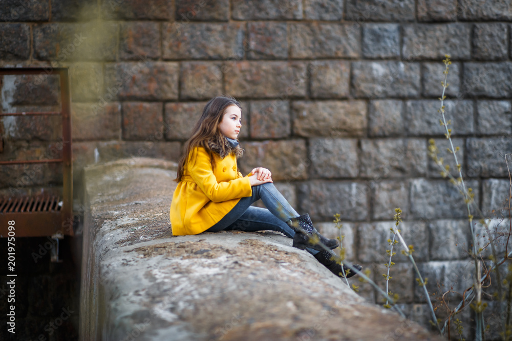 A little girl in a yellow coat under the railway iron bridge at the sunset in a cold evening