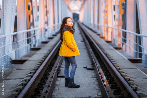 A little girl in a yellow coat walking on the railway iron bridge at the sunset in a cold evening