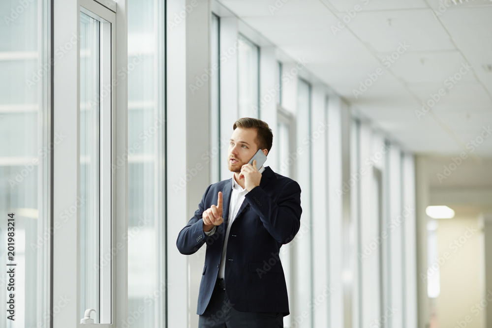 Young businessman standing near the window at office and talking on cell phone Young businessman standing near the window at office and talking on cell phone