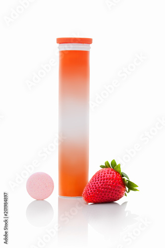 Tube of sparkling tablets with one piece next to tube. Pipe with fizzy tablets with vitamin c. Pastils with straberry flavour with fresh fruit next to. Healing tabletes in tube and one piece outside photo